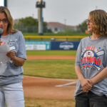 Hadley stands in front of a baseball field and beside a female speaker with a microphone at the Duel on the Diamond event