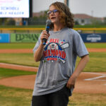 Hadley holds a microphone in front of a baseball field and speaks to others at the Duel on the Diamond event