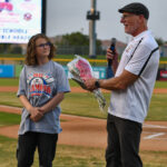 Hadley stands in front of a baseball field and beside a male speaker with a microphone in one hand and a bouquet of red roses in another at the Duel on the Diamond event