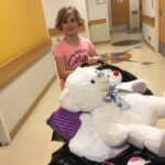 Hadley standing with a large teddy bear in a hospital hallway.