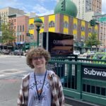 Hadley smiles outside a subway station with a SparkCon lanyard around their neck.