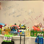 A close shot of a table full of donated cleaning supplies for the Hadley's Bags of Hope event. Behind the supplies are several rose gold balloons and a whiteboard that reads "Welcome to Maricopa West Regional Office! Thank you, Hadley!