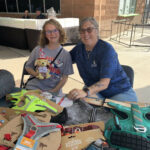 Hadley holds stuffed dog behind a table of pet donations, with a smiling woman next to her.