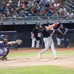 Baseball player takes a swing at a ball beside the umpire in front of an onlooking crowd in the stands at the Duel on the Diamond event