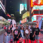 Hadley standing with teenagers on a red carpet, smiling at the camera, in a city at night.