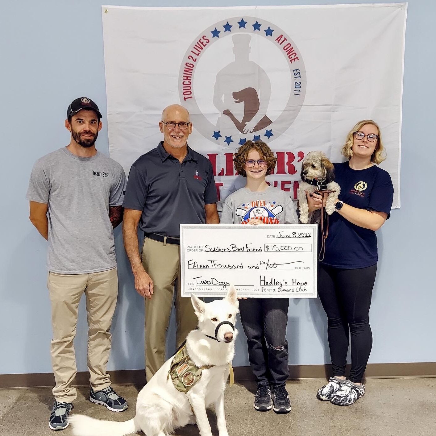 Hadley stands with veterans and two dogs and holds a large novelty check for $15,000 made out to "Soldier's Best Friend" organization.