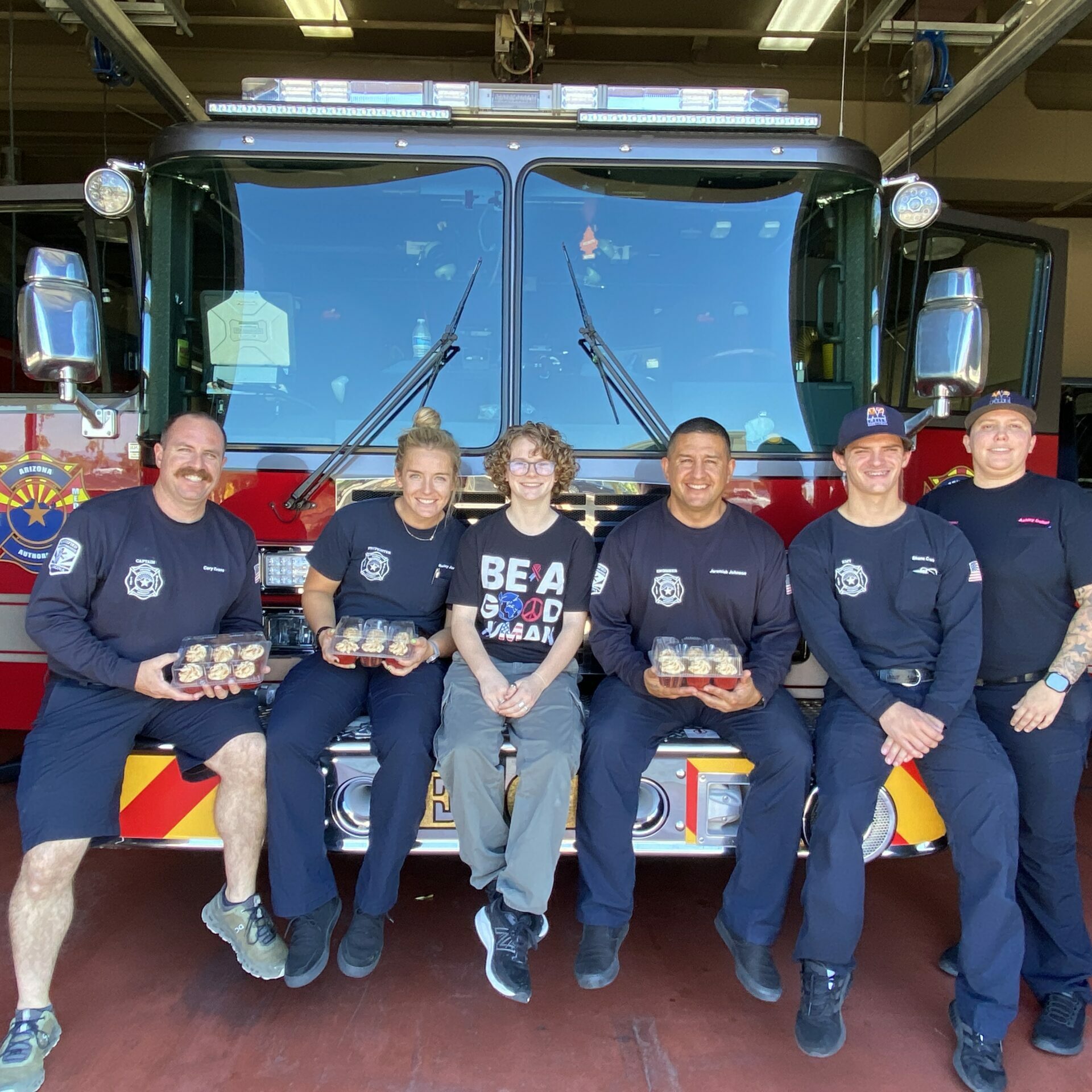 Hadley sits on the front bumper of a firetruck with five firefighters holding cupcakes and smiling.